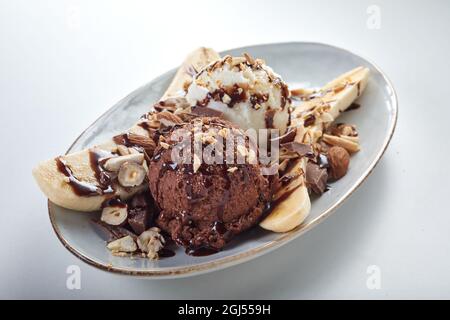 High angle of cold ice cream balls placed between banana slices garnished with chocolate syrup and hazelnuts on white background Stock Photo