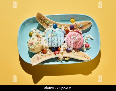 Top view of delicious banana split dessert with colorful ice cream scoops decorated with dragee and sprinkles served on blue plate on yellow backgroun Stock Photo