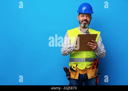 Male builder or handyman in uniform holding clipboard against blue background Stock Photo