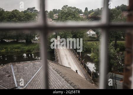 Looking out of old vintage pane glass window from high up angle at old stone bride crossing wide fast flowing river in countryside. Stock Photo