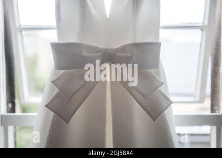 Beautiful pretty big bow on front of brides bright elegant white wedding dress hanging up in the window of the bridal suite on the morning ceremony Stock Photo