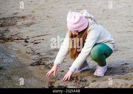 A little girl sits on the banks of the river and touches the surface of the cold water with her own hands. Little girl plays on the river bank. She to Stock Photo
