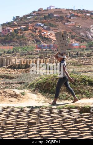 Africa, Madagascar, Antananarivo Province. Brick making factory, a young Malagasy girl carrying dried bricks on her head to where they will be fired. Stock Photo