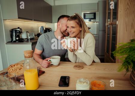 Caucasian happy couple sitting at kitchen table whispering in ear  Stock Photo