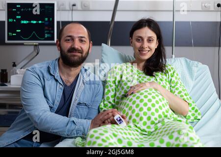 Caucasian people with pregnancy looking at camera and sitting in hospital ward. Smiling couple expecting child and baby delivery while pregnant woman laying in bed at medical clinic Stock Photo