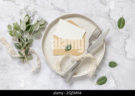 Blank paper card in envelope laying on white plate with fork and knife on marble table with eucalyptus branches and vintage ribbons around, top view. Stock Photo