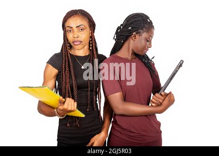 two beautiful female students standing with braids on a white background holding a laptop and book after class. Stock Photo