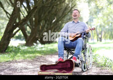Disabled man in wheelchair plays guitar in park and makes money Stock Photo