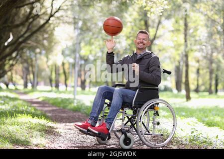Smiling disabled man twirls basketball ball on his finger while sitting in wheelchair in park Stock Photo