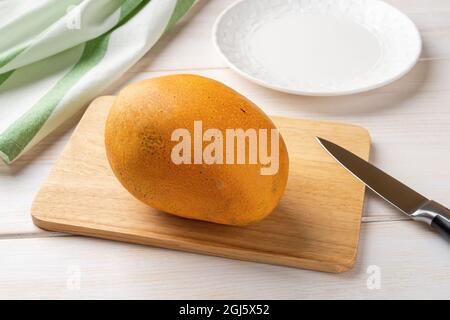 Very ripe whole mango and fruit knife on a cutting board over white wooden table. Fresh yellow delicious mango. Ready to eat sweet tropical fruits. Stock Photo