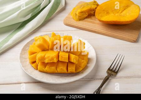Cut ripe juicy mango on a white plate and fruit fork over wooden table. Yellow delicious mango flesh sliced with cubes. Sweet tropical fruits. Stock Photo