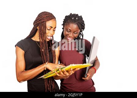 Two beautiful female students standing on a white background looking at a magazine and laughing while holding a laptop. Stock Photo