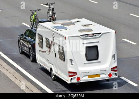 Arial side & back view of white Swift Challenger 590 towing holiday caravan roof solar panel & towed by black car with bikes on roof rack UK motorway Stock Photo