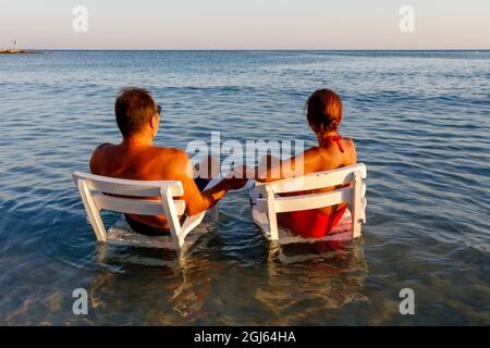 Couple holding hands sitting on armchairs in the water, beach in Cesme, Izmir, Turkey. Stock Photo