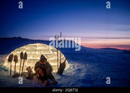 Baker Lake, Nunavut, Canada. Man in traditional caribou skin clothing, silhouetted with Inuit tools against a lit igloo at dusk. (MR) Stock Photo