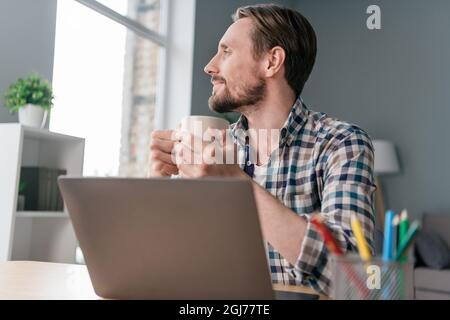 Photo of positive dreamy mature guy dressed checkered shirt sitting table working modern device drinking tea indoors apartment office Stock Photo