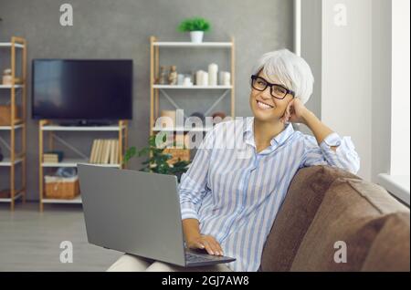 Portrait of happy mature woman sitting on sofa at home, holding laptop and smiling Stock Photo