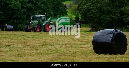 Hay or silage making (farmer in farm tractor at work in rural field pulling baler, collecting dry grass & round bales wrapped) - Yorkshire England UK. Stock Photo