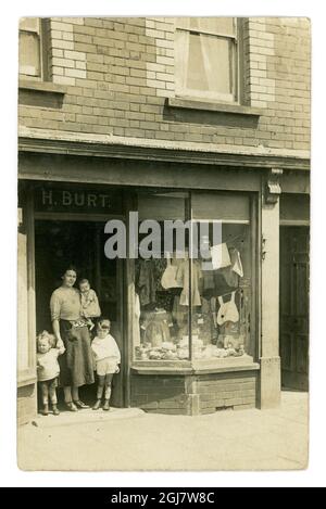 Original real photographic postcard of working class woman with her children standing outside in shop doorway of a haberdashery store, called H. Burt, selling wool, aprons, camisoles, wool and threads. The woman looks careworn and the shopfront is shabby, probably in a poor neighbourhood, circa 1921, UK Stock Photo