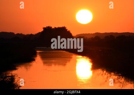 Beautiful sun reflecting over calm water with distant trees and orange sky Stock Photo