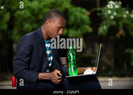 young african man sitting in a park and working on his laptop computer with a plastic water bottle laying on it Stock Photo