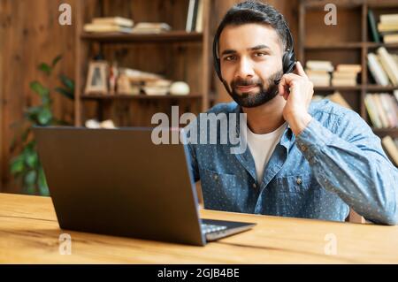 Portrait of confident Indian guy in wireless headset using laptop computer sitting in contemporary loft style office space, man is making video call, talking online on distance Stock Photo