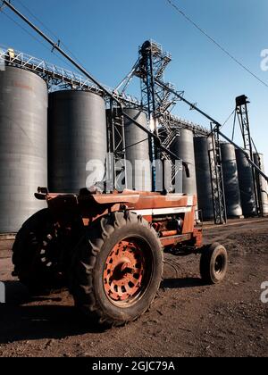 Old red work tractor parked in front of several large steel grain silos on farm Stock Photo