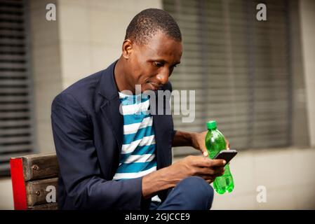 Thoughtful young man texting on the phone and drinking juice while sitting on a bench near a modern office building Stock Photo