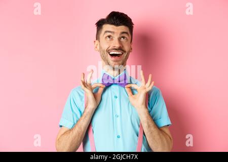 Happy young man with moustache, touching his bow-tie and laughing, looking up at logo, standing on pink background Stock Photo