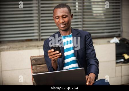 Image of a beautiful young happy businessman sitting in the street on a bench using a mobile phone and holding a laptop while smiling Stock Photo