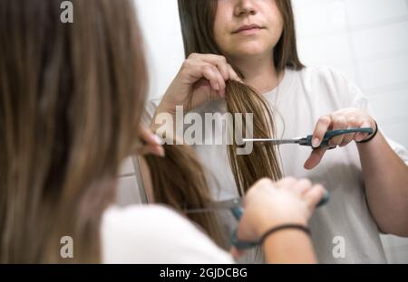 Young woman in home bathroom looking in mirror after cutting her own hair  with scissors Stock Photo - Alamy