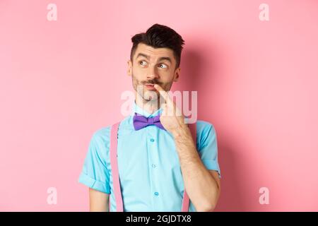 Image of handsome funny man in bow-tie looking thoughtful left and touching lip, thinking what to choose, making decision, standing over pink Stock Photo