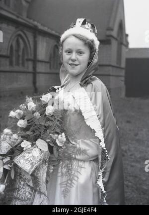 1956, historical, a teenage girl, the May Day Queen standing in the grounds of a church wearing her crown, gown, robe and holding a bouquet of flowers before taking part in the traditional May Day carnival, Leeds, England, UK. Stock Photo