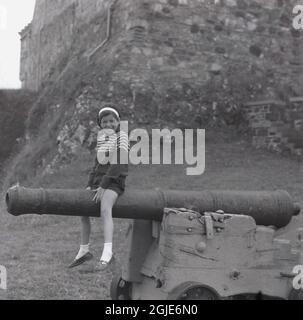 1961, historical, in the Scottish highlands, a young girl sitting aside an old cannon mounted on a wooden wheeled support in the grounds below Duart Castle, Inner Hebrides, Isle of Mull, Scotland, UK. Stock Photo