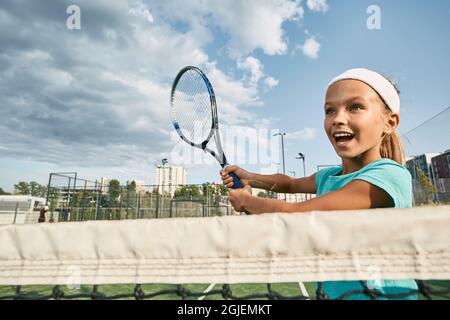 Beautiful female child tennis player playing forehand near net while tennis match on city court outdoors, low angle view Stock Photo