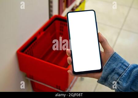 Mockup image of woman hand holding blank white desktop screen mobile phone in the supermarket. Blurry plastic red shopping baskets in the background. Stock Photo