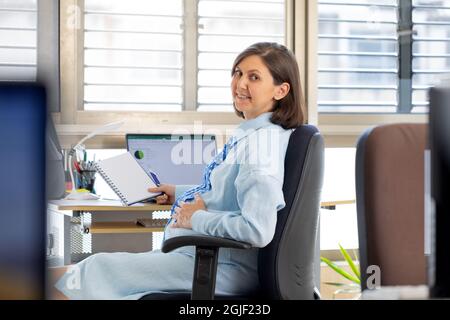 pregnant woman works in the office at the computer. The employee sits on a chair opposite the window and turns.  Stock Photo