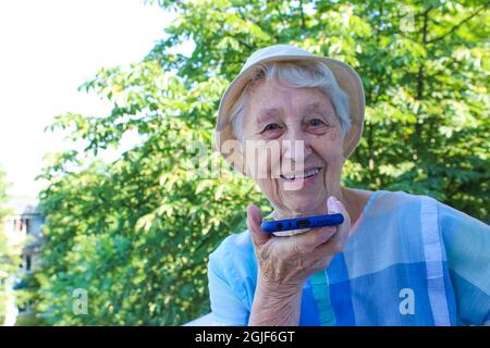 Charming elderly woman in rectangular black rimmed eyewear using voice recognition function on cell phone. Modern senior female holding electronic gadget, sending vocal message via online messenger Stock Photo