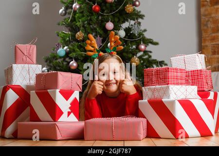 The child lies on the floor surrounded by boxes with gifts against the background of a Christmas tree. Little girl in a deer headband showing thumb up Stock Photo