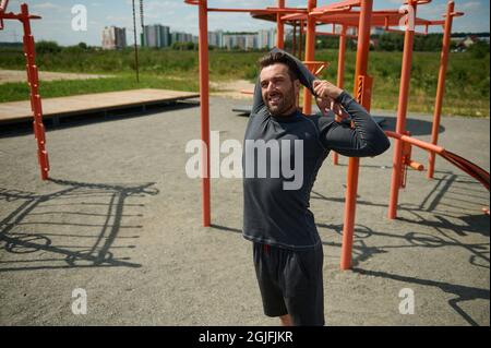 Handsome mature sportsman stretching arms behind his back before training on the sportsground in open air. Young adult 40 years old European sporty ma