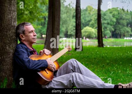 In summer evening, adult romantic man sits in beautiful park on grass, leaning against tree, and plays six-string classical acoustic guitar. Selective Stock Photo