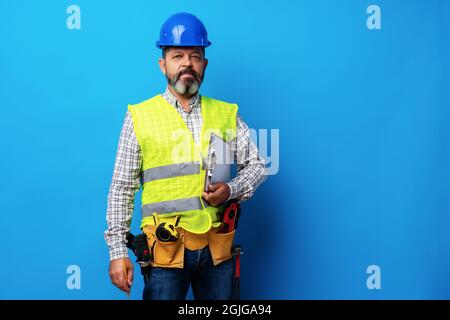 Male builder or handyman in uniform holding clipboard against blue background Stock Photo