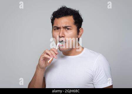 Mexican man with Latino Hispanic appearance brushes his teeth with an orange toothbrush, young man with Chinese hair in white shirt with gray backgrou Stock Photo