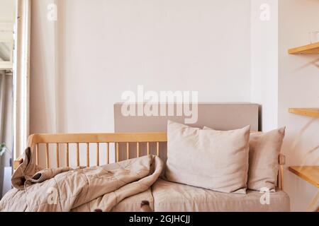 Fragment of home interior in natural pastel tones with wooden couch with soft beige cushions and coverlet placed against white wall Stock Photo