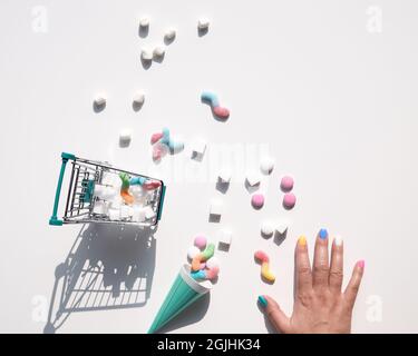 Shopping cart with shugar cubes. Hand and various tasty snacks in pastel colors, pink, mint green, on white background. Excess of sweets, chocolate Stock Photo