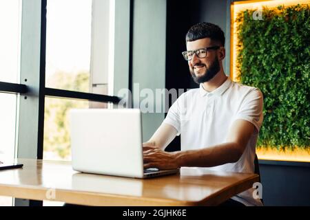 Happy young hipster wearing glasses and a white t-shirt using a laptop while sitting at a table in a cafe, Young man who is working on a laptop at the Stock Photo