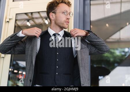 curly businessman in glasses and classy suit wearing blazer near cafe entrance Stock Photo