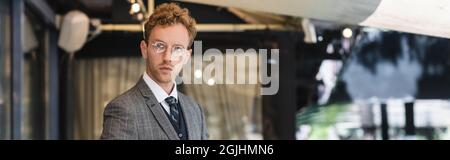 curly businessman in glasses and classy suit near cafe entrance, banner Stock Photo