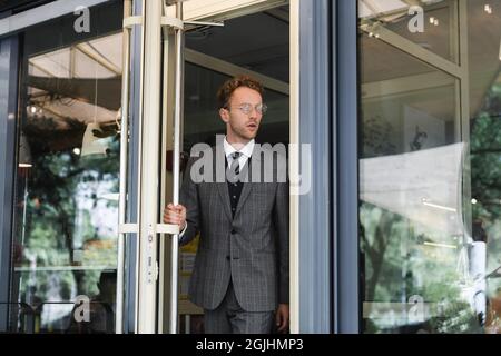 curly businessman in glasses opening door while leaving cafe Stock Photo