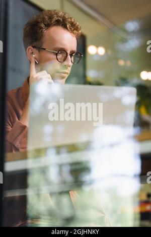 young man in glasses adjusting earphones near laptop behind blurred window of cafe Stock Photo